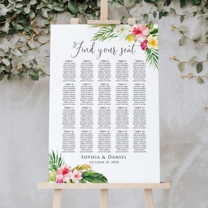 May include: A white wedding seating chart on an easel, featuring the words "Find your seat" in elegant script. The chart lists guest names under table numbers, with floral and leaf accents in pink, white, and green. The names Sophia & Daniel and the date are at the bottom.