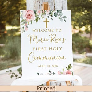 May include: A white sign on an easel with gold text and floral accents. The sign reads "Welcome to Maria Rose's First Holy Communion" with the date April 20, 20XX. The top and bottom of the sign are decorated with pink roses and green leaves.