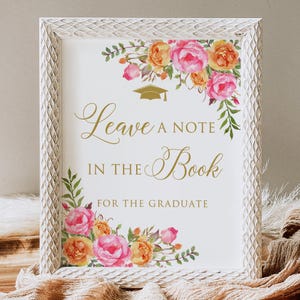 May include: A framed sign with the text "Leave a Note in the Book for the Graduate." The white frame has a textured pattern. Floral arrangements with pink and orange roses and greenery adorn the top and bottom corners. A gold graduation cap is above the text.