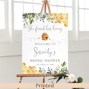 May include: A white bridal shower sign with a beehive and honeycomb design. The text reads "She found her honey, Welcome to Serenity's Bridal Shower, October 25, 20XX".