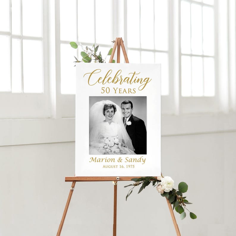 May include: A white sign with gold text celebrating 50 years of marriage. The sign features a black and white photo of a couple on their wedding day. The text reads "Celebrating 50 Years" and "Marion & Sandy August 16, 1975".