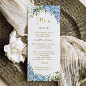 May include: A rectangular menu card with a floral design featuring blue hydrangeas and green leaves. The menu includes a starter, main course, and dessert options, with text in gold script. The card is set on a wooden surface.