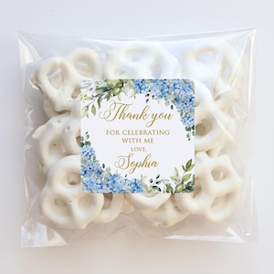 May include: A clear cellophane bag filled with white chocolate-covered pretzels. A square label with blue hydrangea flowers and the text "Thank you for celebrating with me, love, Sophia" is affixed to the bag.
