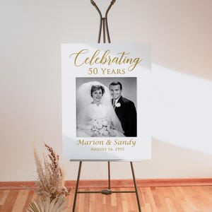 May include: White sign with gold text celebrating 50 years of marriage. A black and white photo of a couple on their wedding day is in the center of the sign. The text below the photo reads "Marion & Sandy August 16, 1975".