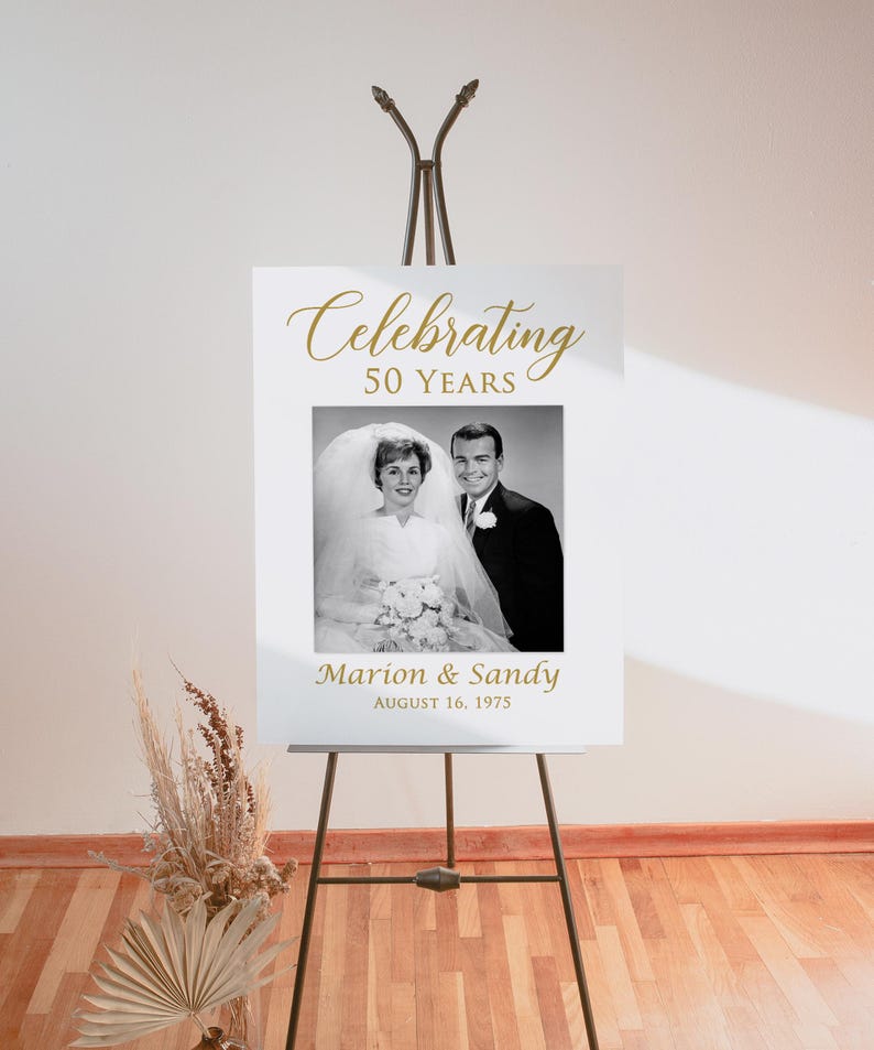 May include: White sign with gold text celebrating 50 years of marriage. A black and white photo of a couple on their wedding day is in the center of the sign. The text below the photo reads "Marion & Sandy August 16, 1975".