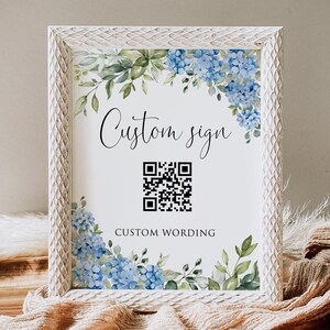 May include: A rectangular sign in a textured white frame featuring blue hydrangea flowers and green leaves at the top and bottom corners. The center displays the text Custom sign in script, a black and white QR code, and the text CUSTOM WORDING.
