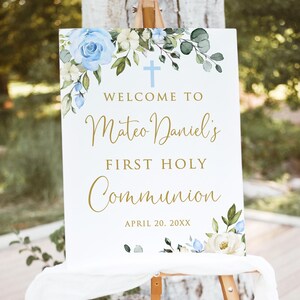 May include: A white sign on an easel with floral accents and text. The sign reads "Welcome to Mateo Daniel's First Holy Communion" with a light blue cross and flowers. The date "April 20, 20XX" is at the bottom.