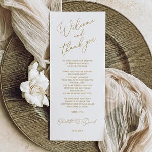 May include: A wedding welcome card with gold script on a white background, featuring the words "Welcome and Thank You." The card is on a decorative plate with a white flower and draped fabric.
