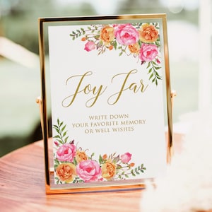 Peut inclure: Panneau doré avec l'inscription "Joy Jar" en lettres dorées, orné de compositions florales à l'aquarelle dans les tons rose, orange et vert. Le texte en dessous indique "Écrivez votre souvenir préféré ou vos bons vœux."