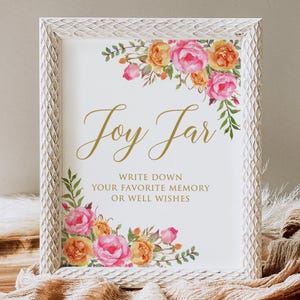 May include: A framed sign with the words "Joy Jar" in gold script, surrounded by watercolor floral arrangements in pink, orange, and yellow. The sign encourages guests to write down memories or well wishes. The frame is white with a textured pattern.