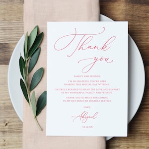 May include: A white card with the words "Thank you" in a cursive, red font. The card includes a message of gratitude and is placed on a white plate with a sprig of green leaves. The card is for a Bat Mitzvah.
