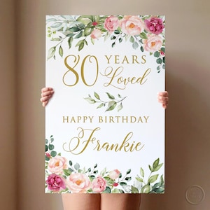 May include: A white sign with pink and green floral borders. The sign says "80 Years Loved" in gold lettering and "Happy Birthday Frankie" in gold cursive lettering.