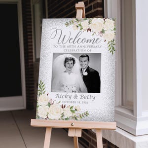 May include: A welcome sign for a 60th-anniversary celebration. The sign features a black and white photo of a couple, with the names "Ricky & Betty" and the date "October 18, 1956" below. The sign is decorated with white roses and greenery.