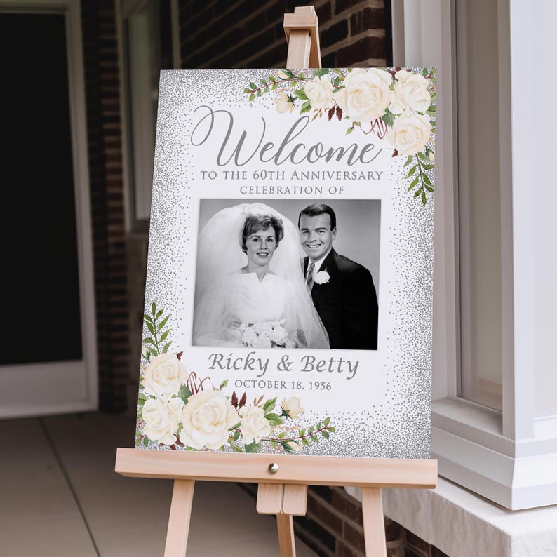 May include: A welcome sign for a 60th-anniversary celebration. The sign features a black and white photo of a couple, with the names "Ricky & Betty" and the date "October 18, 1956" below. The sign is decorated with white roses and greenery.