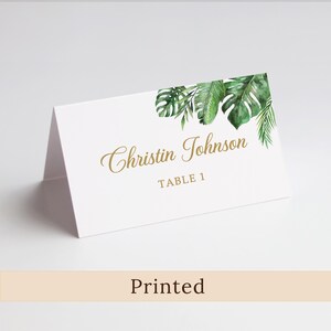 May include: White folded place card with gold script text reading "Christin Johnson" and "TABLE 1." The upper right corner features a watercolor design of green tropical leaves. The word "Printed" is at the bottom.