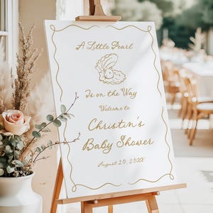 May include: A white sign on a wooden easel with gold lettering and a decorative border. The sign reads "A Little Pearl Is on the Way Welcome to Christin's Baby Shower August 20, 20XX" with an illustration of an oyster.