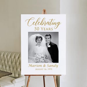 May include: White sign with gold text celebrating 50 years of marriage. The sign features a black and white photo of a couple on their wedding day. The text reads "Celebrating 50 Years" and "Marion & Sandy August 16, 1975".