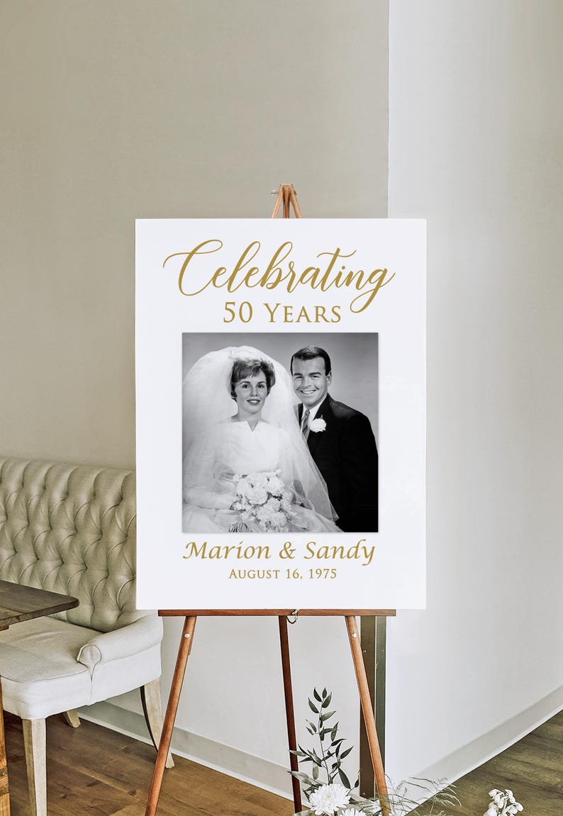 May include: White sign with gold text celebrating 50 years of marriage. The sign features a black and white photo of a couple on their wedding day. The text reads "Celebrating 50 Years" and "Marion & Sandy August 16, 1975".