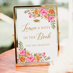 May include: A gold framed sign with a white background and floral design. The sign says "Leave a note in the book for the graduate".