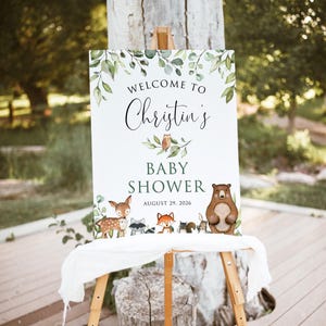 May include: Welcome to Christin's Baby Shower sign with a woodland theme. The sign features a green leafy border with a brown bear, a deer, a raccoon, a fox, and an owl. The text on the sign reads "Welcome to Christin's Baby Shower August 29, 2026".