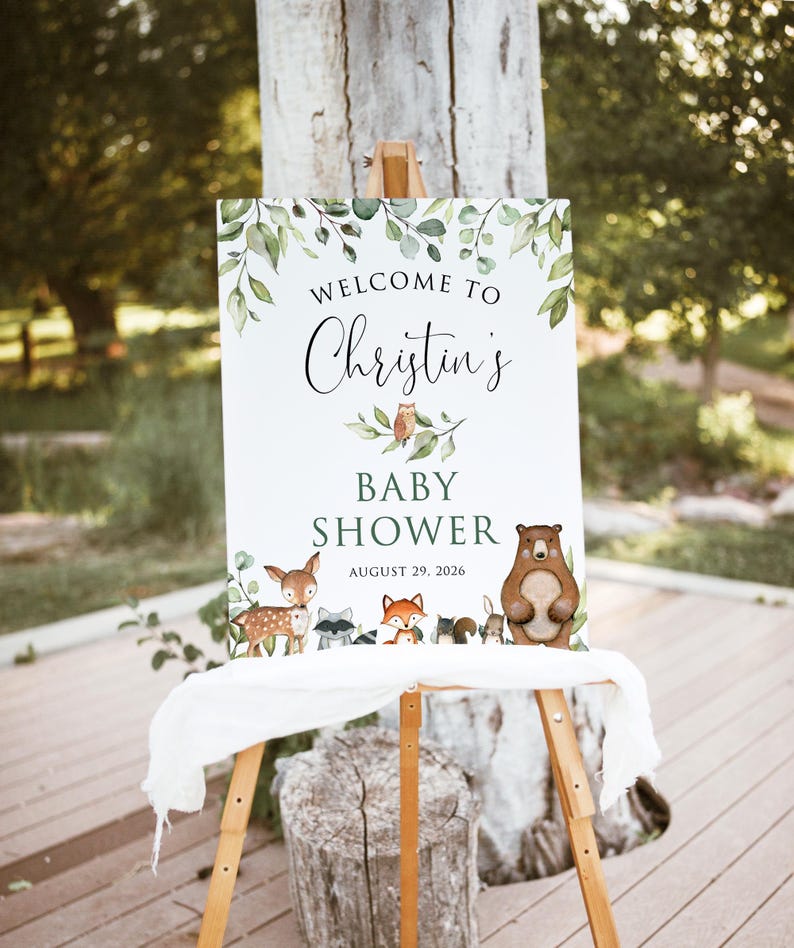 May include: Welcome to Christin's Baby Shower sign with a woodland theme. The sign features a green leafy border with a brown bear, a deer, a raccoon, a fox, and an owl. The text on the sign reads "Welcome to Christin's Baby Shower August 29, 2026".