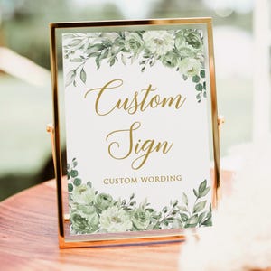 Peut inclure: Un panneau doré avec l'inscription "Custom Sign" en lettres dorées élégantes. Le panneau est orné de compositions florales à l'aquarelle dans des tons verts et blancs. Les mots "Custom Wording" sont en dessous.