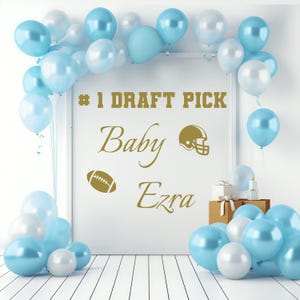 May include: A white framed sign with blue and white balloons around it. The sign says "#1 Draft Pick Baby Ezra" with a football and a football helmet.