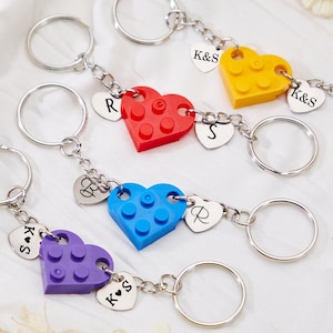 May include: Four silver keychains with heart-shaped charms and small Lego-like heart-shaped charms in red, yellow, blue, and purple. The charms have initials on them, including K&S, R, and S.