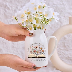 Puede incluir: Un jarrón de cerámica blanco con asa, lleno de flores blancas. El jarrón presenta un diseño floral con las palabras "teach them, love them, watch them grow" y el nombre "mrswilson".