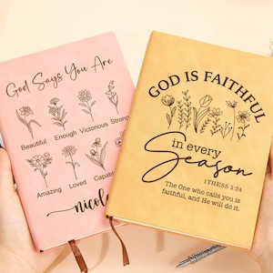 Pode incluir: Dois diários rosa e bege com designs florais e citações inspiradoras. O diário rosa diz "Deus diz que você é" com uma lista de afirmações positivas. O diário bege diz "Deus é fiel em todas as estações" com um versículo bíblico de 1 Tessalonicenses 5:24.