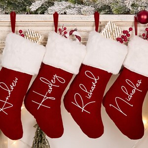 Könnte beinhalten: Vier rote Weihnachtsstrümpfe mit weißem Kunstfellbesatz und weißen personalisierten Namen: Andrew, Hanley, Richard und Nicholas.