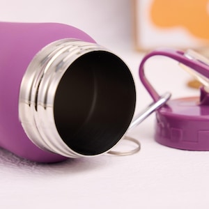 May include: A close-up of a purple water bottle with a stainless steel rim and a matching purple lid. The lid has a flip-top design and a silver handle. The bottle is lying on a white surface.