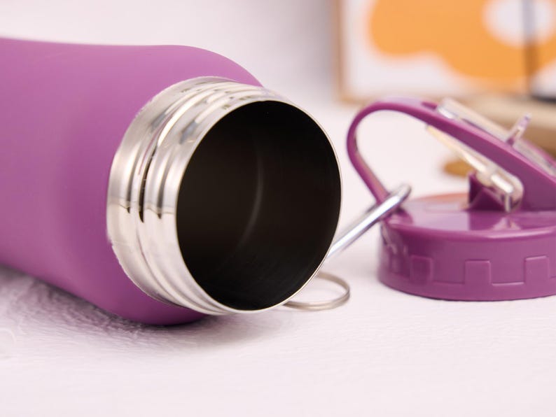 May include: A close-up of a purple water bottle with a stainless steel rim and a matching purple lid. The lid has a flip-top design and a silver handle. The bottle is lying on a white surface.