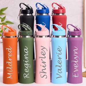 May include: A collection of insulated water bottles in various colors, including orange, green, pink, blue, and purple. Each bottle has a name printed on it in a cursive font. The bottles have a flip-top lid and a silver carabiner clip.
