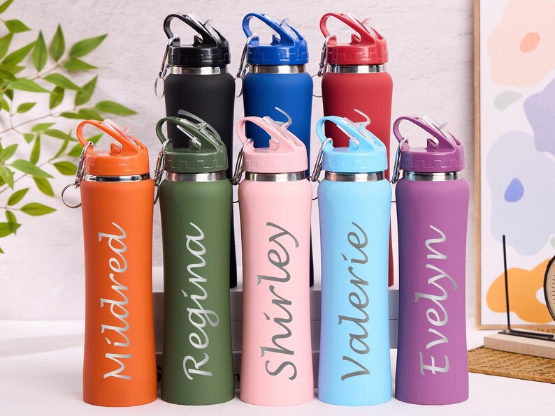 May include: A collection of insulated water bottles in various colors, including orange, green, pink, blue, and purple. Each bottle has a name printed on it in a cursive font. The bottles have a flip-top lid and a silver carabiner clip.