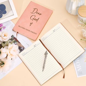 May include: Two notebooks, one pink with the text "Dear God" and "Prayers from Jordan" and the other with lined pages, on a light beige surface with a bouquet of white flowers.