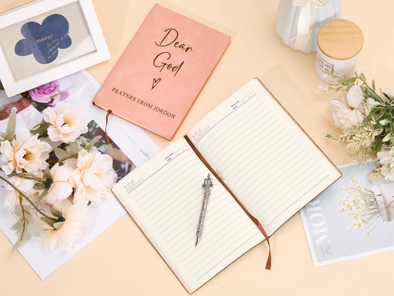 May include: Two notebooks, one pink with the text "Dear God" and "Prayers from Jordan" and the other with lined pages, on a light beige surface with a bouquet of white flowers.