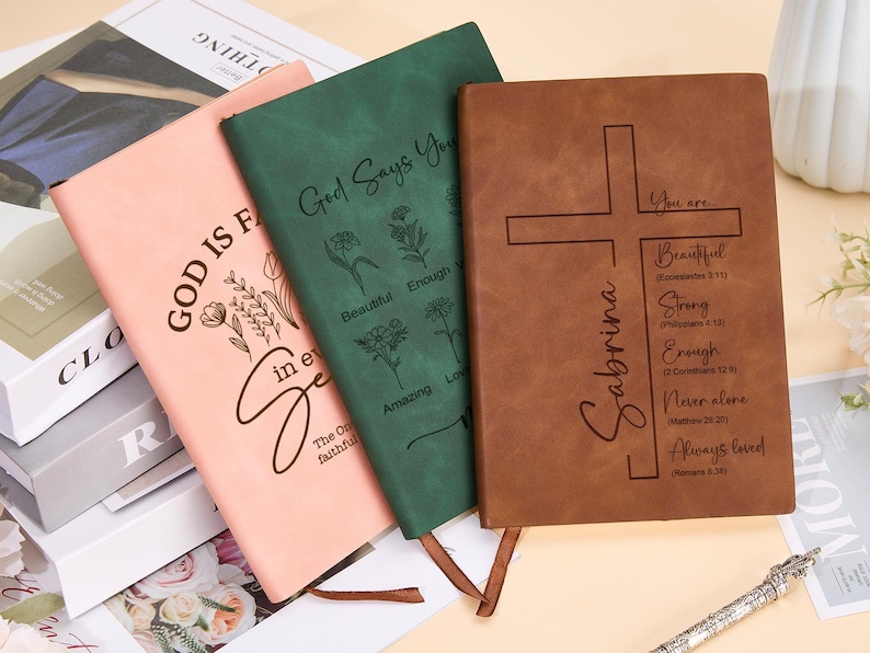 May include: Three leather-bound journals with inspirational quotes and floral designs. The pink journal says "God is faithful in every season". The green journal says "God says you are beautiful, enough, amazing, loved". The brown journal says "Sabrina" and has a cross with the words "You are beautiful, strong, enough, never alone, always loved".