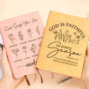 May include: Two pink and beige leather-bound journals with inspirational quotes and floral designs. The pink journal says "God Says You Are" with a list of positive affirmations. The beige journal says "God is Faithful in Every Season" with a Bible verse from 1 Thessalonians 5:24.