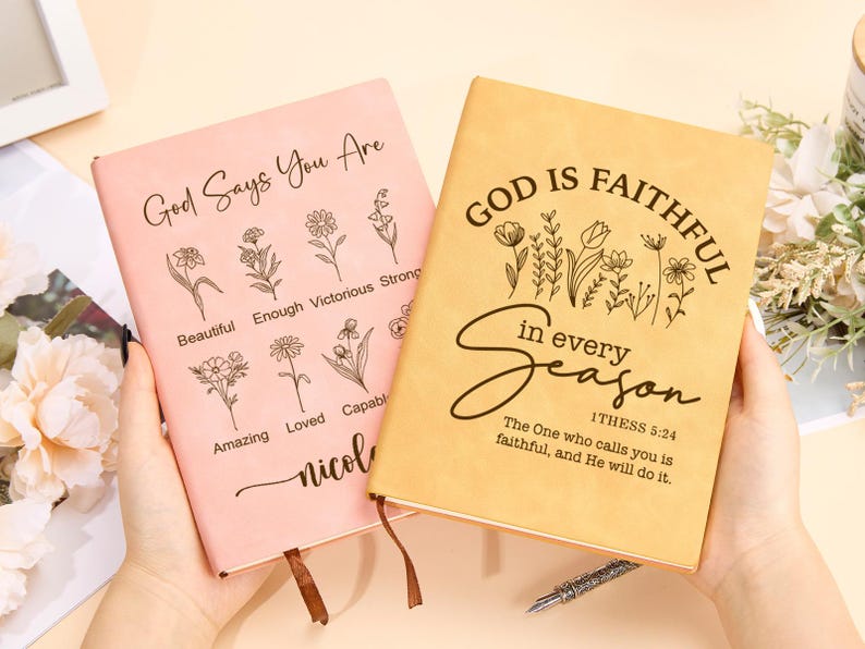 May include: Two pink and beige leather-bound journals with inspirational quotes and floral designs. The pink journal says "God Says You Are" with a list of positive affirmations. The beige journal says "God is Faithful in Every Season" with a Bible verse from 1 Thessalonians 5:24.