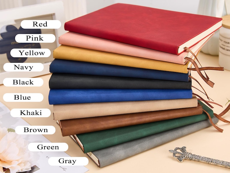 May include: A stack of ten notebooks in various colors, including red, pink, yellow, navy, black, blue, khaki, brown, green, and gray. The notebooks are made of leather and have a simple design.