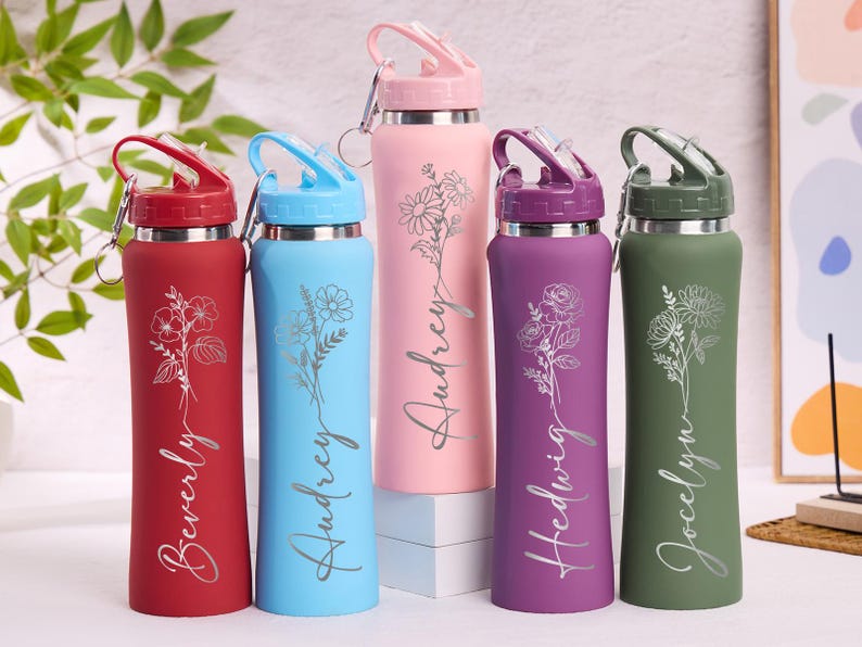 May include: A collection of insulated water bottles in various colors: red, blue, pink, purple, and green. Each bottle features a floral design and a name in a cursive font. The bottles have a flip-top lid and a silver carabiner clip.