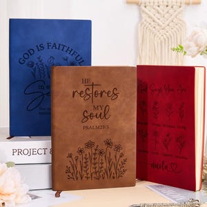 May include: Three leather-bound journals with different inspirational quotes and floral designs. The first journal is blue with the text "God is Faithful in Every Situation". The second journal is brown with the text "He Restores My Soul Psalm 23:3". The third journal is red with the text "God Says You Are" and a floral design.