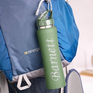 May include: A green water bottle with the name "Barnett" printed vertically in white. The bottle is attached to a blue backpack with a carabiner. The bottle has a green lid and a silver rim.