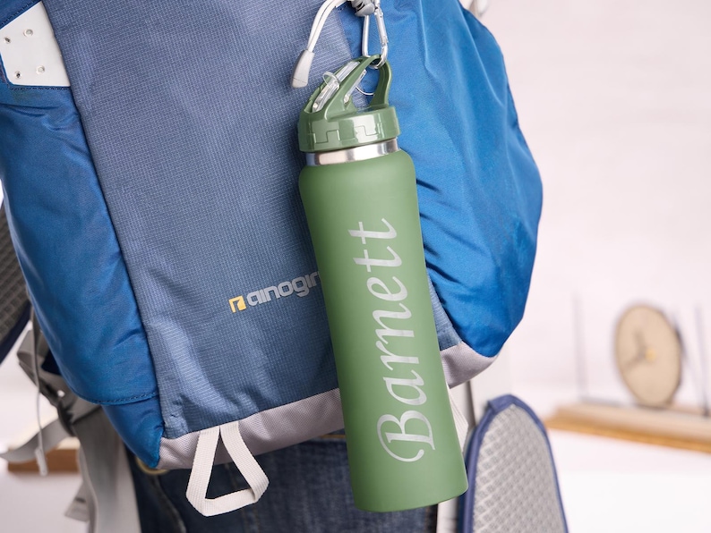 May include: A green water bottle with the name "Barnett" printed vertically in white. The bottle is attached to a blue backpack with a carabiner. The bottle has a green lid and a silver rim.