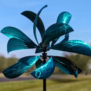 Könnte beinhalten: Ein teal-farbener Metall-Windspinner mit gebogenen, blütenblattförmigen Flügeln. Die Flügel sind kreisförmig um eine zentrale Nabe angeordnet und auf einer schwarzen Stange montiert. Der Windspinner befindet sich im Freien mit blauem Himmel im Hintergrund.