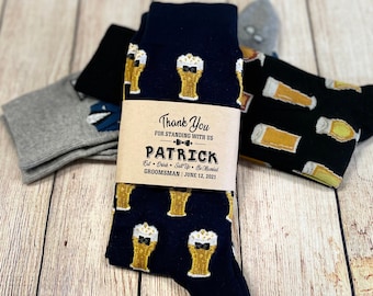 May include: A pair of black socks with a beer mug pattern. The socks have a brown paper tag that says "Thank You for Standing With Us Patrick Eat Drink Salt Up Be Married Groomman June 12, 2021".
