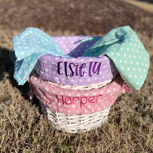 May include: Two white wicker baskets with polka dot fabric liners. The top liner is lavender with the name "Elsie Lu" embroidered in purple. The bottom liner is pink with the name "Harper" embroidered in pink.