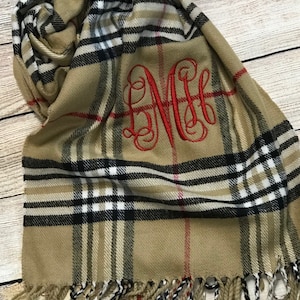 May include: A tan and black plaid scarf with red accents and a red embroidered monogram. The scarf has fringe on the bottom.