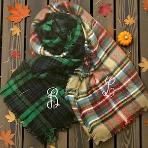 May include: Two plaid scarves in green, black, red, and tan with white embroidered initials. One scarf is folded, and the other is spread out. Autumn leaves and a small pumpkin are scattered around the scarves on a wooden surface.